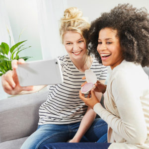 Two young women in a living room holding Caya in a clamshell case and either taking a picture with a cell phone or sharing their image with friends.