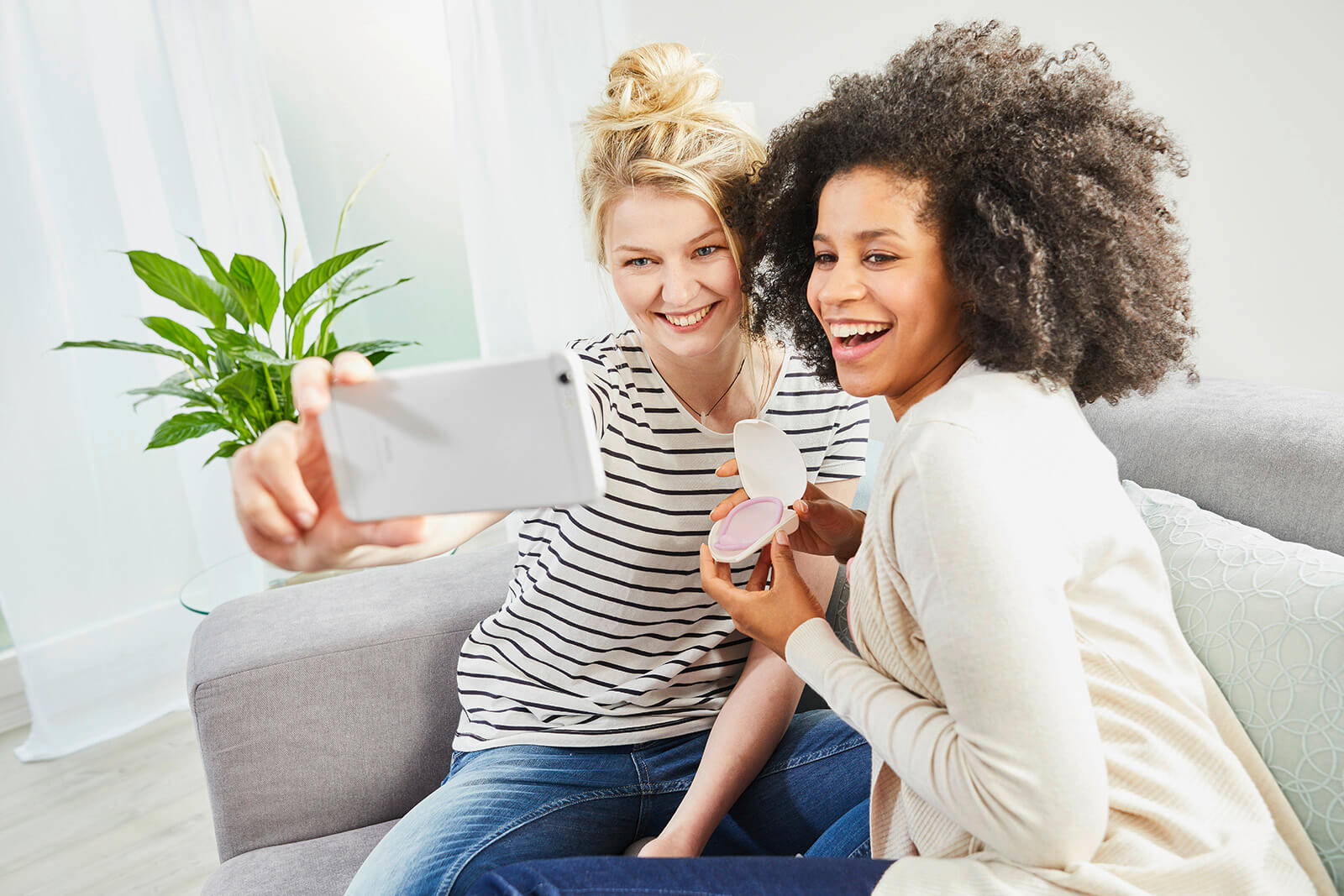 Two young women in a living room holding Caya in a clamshell case and either taking a picture with a cell phone or sharing their image with friends.