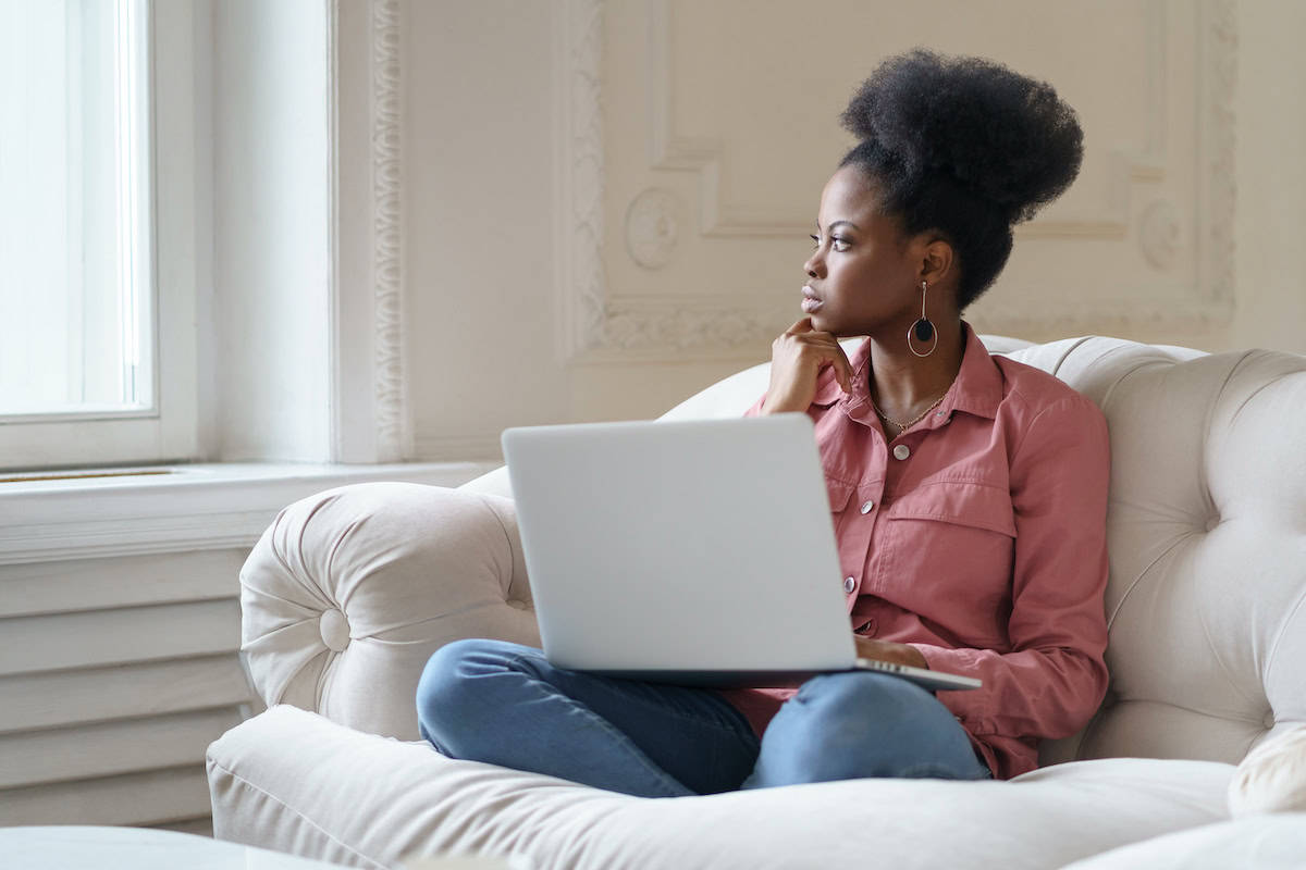 Thoughtful serious African American millennial woman with afro hairstyle wear pink shirt sitting on couch at home, distracted from laptop work looking out the window, thinking about problems.
