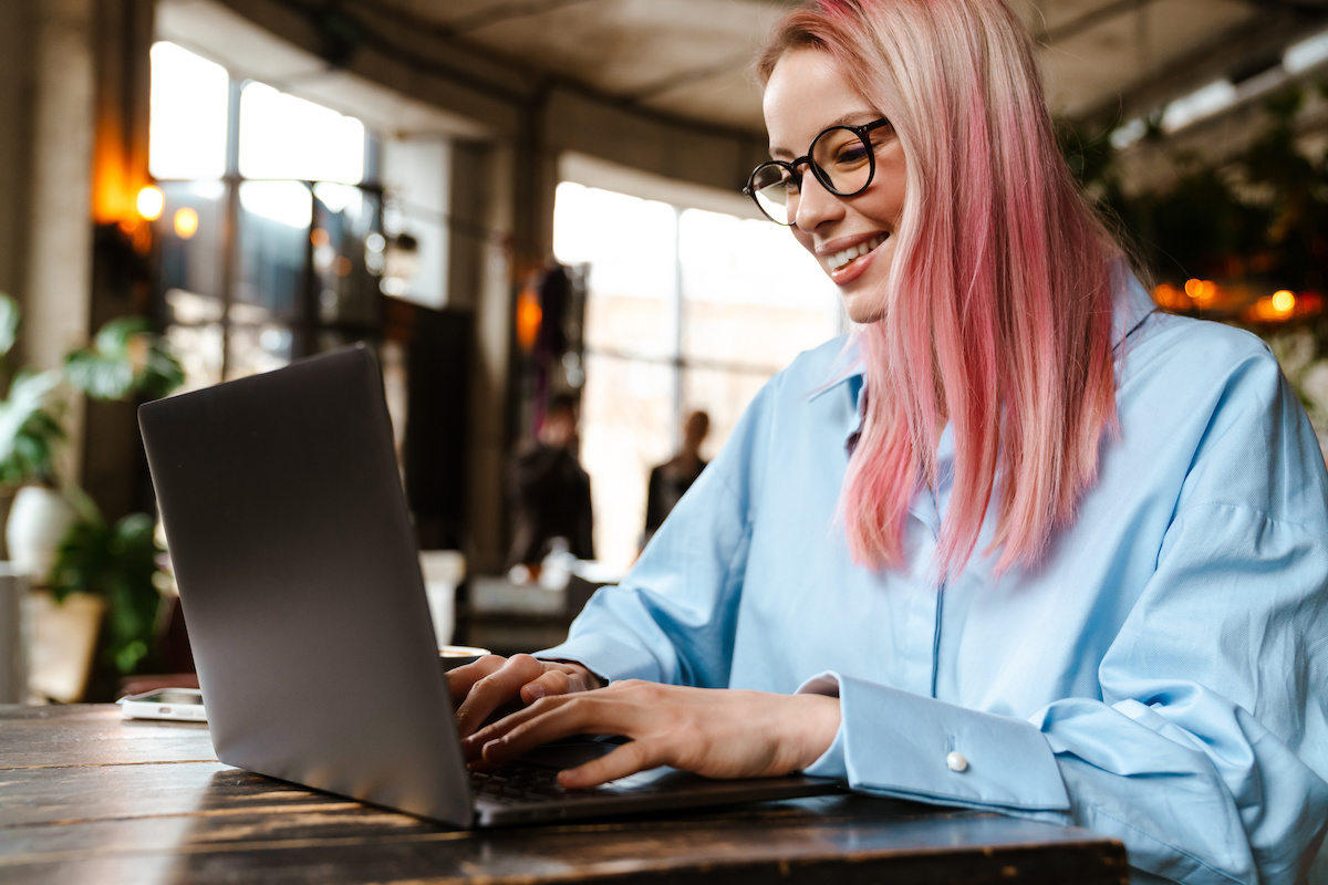 Young beautiful woman smiling and using laptop while sitting in cafe. Caya pink hair.