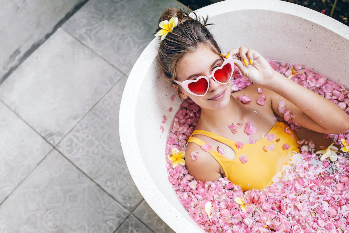 Young teenage girl in funky heart shape sunglasses relaxing in organic bath tube with tropical flowers in luxurious bath. Pink petals like Caya Contraception.