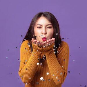 Young Asian woman cheerful blowing confetti on banner isolated purple studio background. Asian female happy in orange sweater.