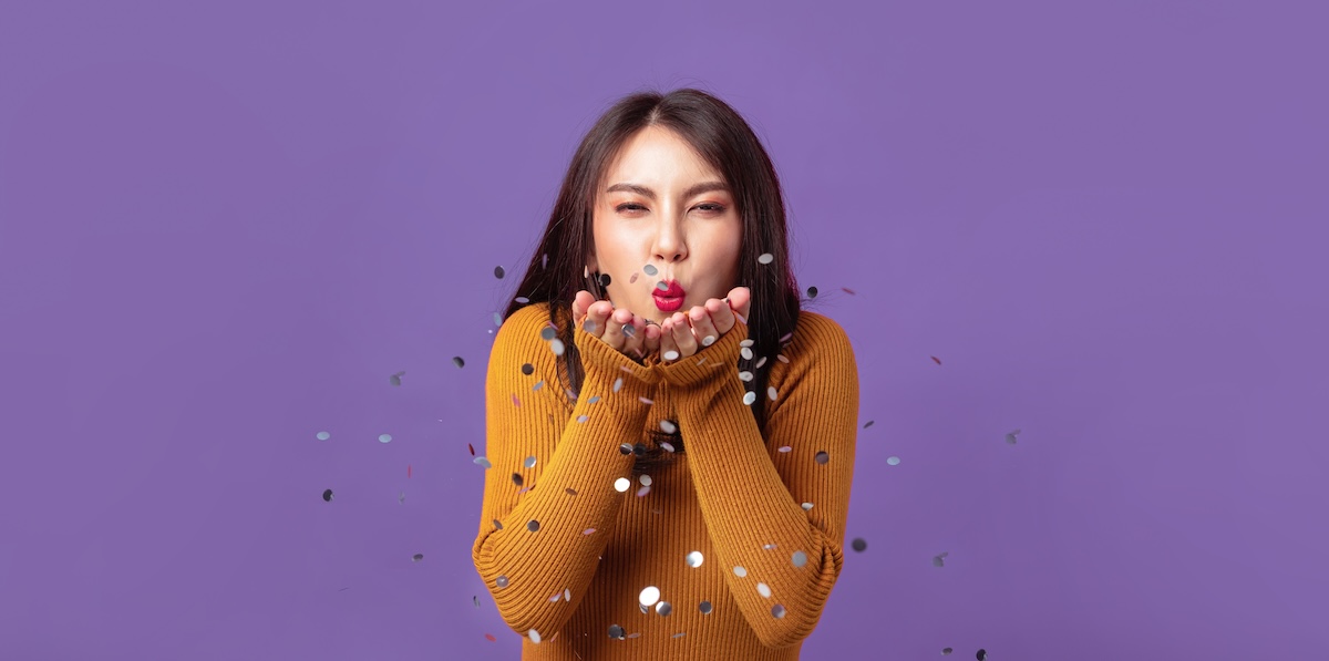 Young Asian woman cheerful blowing confetti on banner isolated purple studio background. Asian female happy in orange sweater.