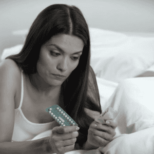 Woman on bed examining birth control pills and condom while her boyfriend is asleep. Contemplating contraception options.
