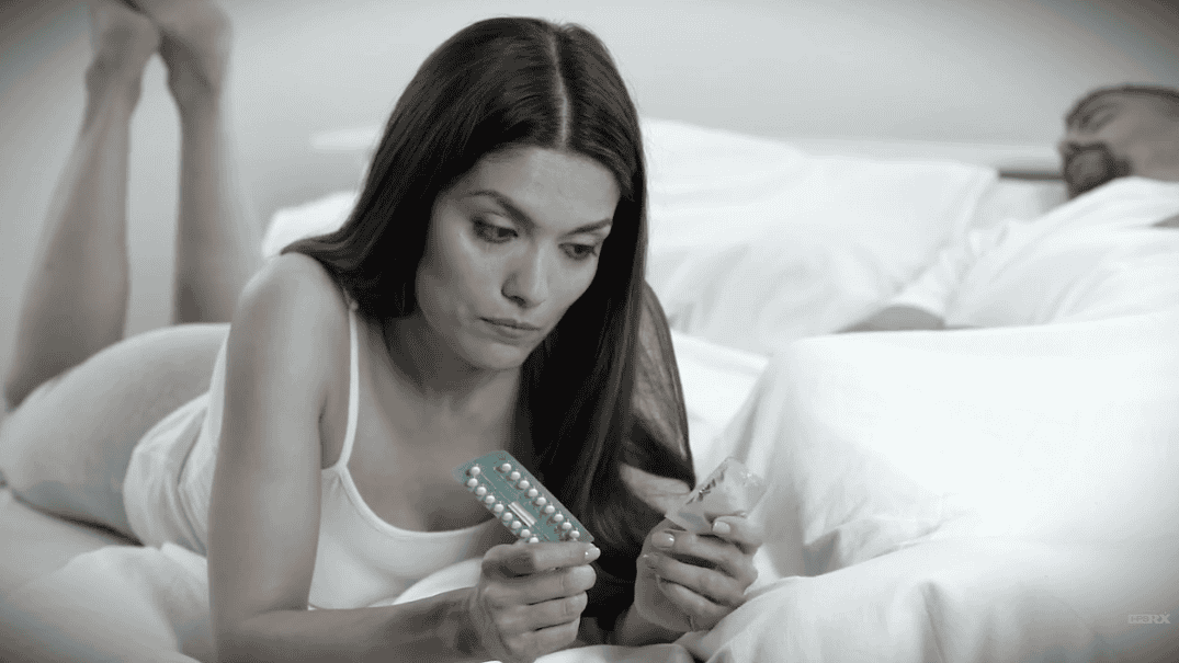 Woman on bed examining birth control pills and condom while her boyfriend is asleep. Contemplating contraception options.