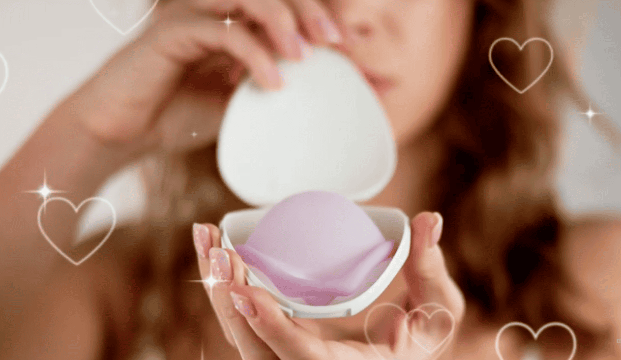 Woman holding a Caya diaphragm inside a clamshell case, close-up, with sparky nails and hearts floating around her.