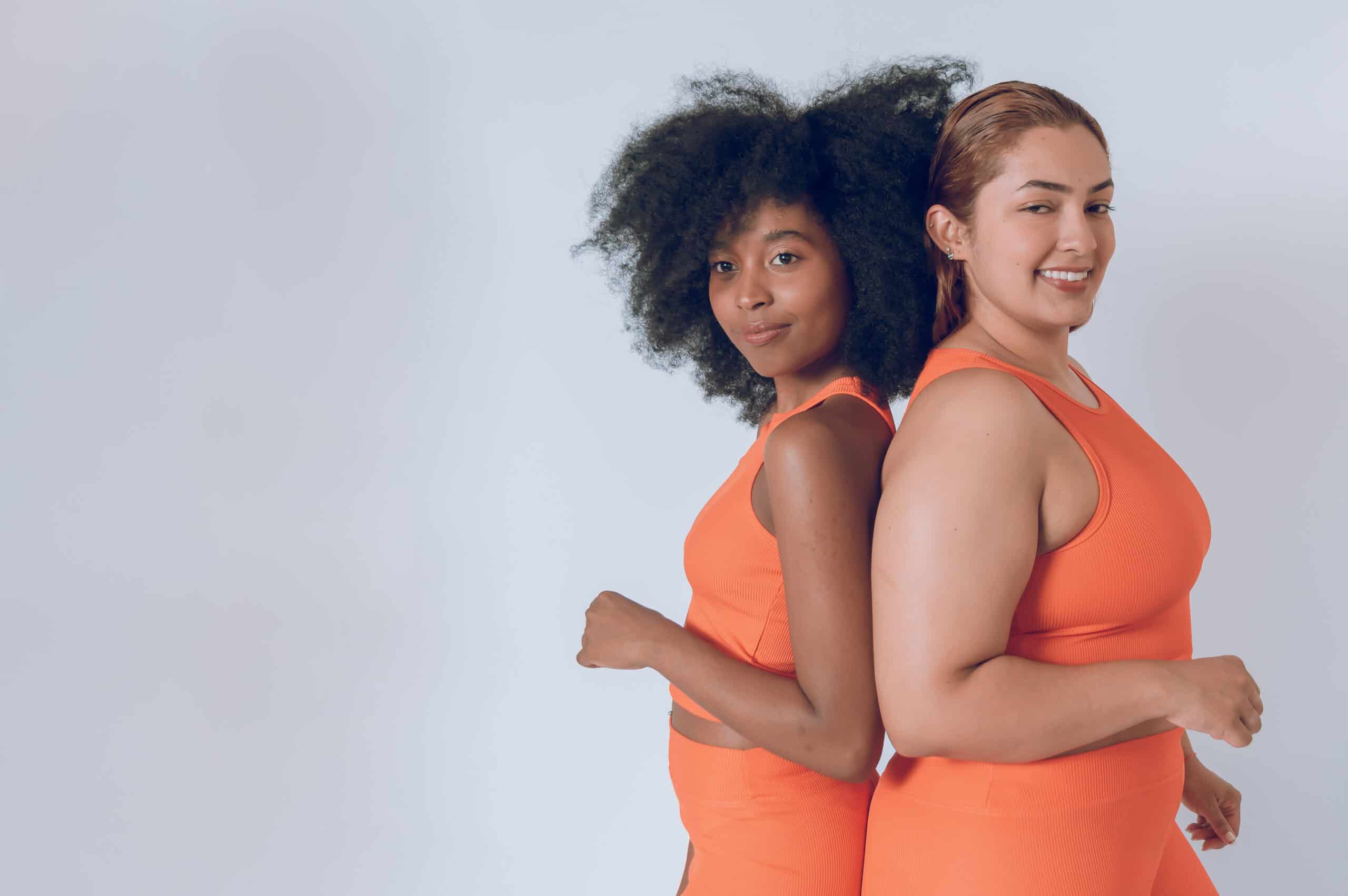 multiethnic beautiful young hispanic latina girls, posing with their backs together, looking at camera, wearing orange sportswear on a white background. image with copy space.