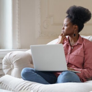 Thoughtful serious African American millennial woman with afro hairstyle wear pink shirt sitting on couch at home, distracted from laptop work looking out the window, thinking about problems.