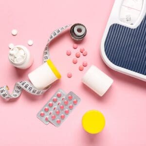 Bottles with weight loss pills, scales and measuring tape on pink background