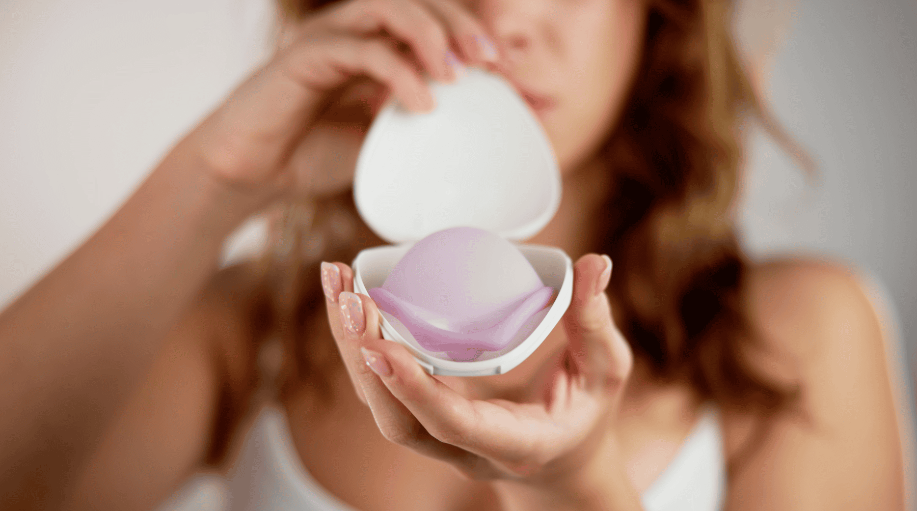 A young woman with long hair holding up a silicone Caya diaphragm that is sitting inside the opened clamshell case.