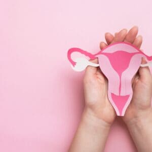 Pink background and hands clasped with a paper cut-out design of a woman's reproduction organs.